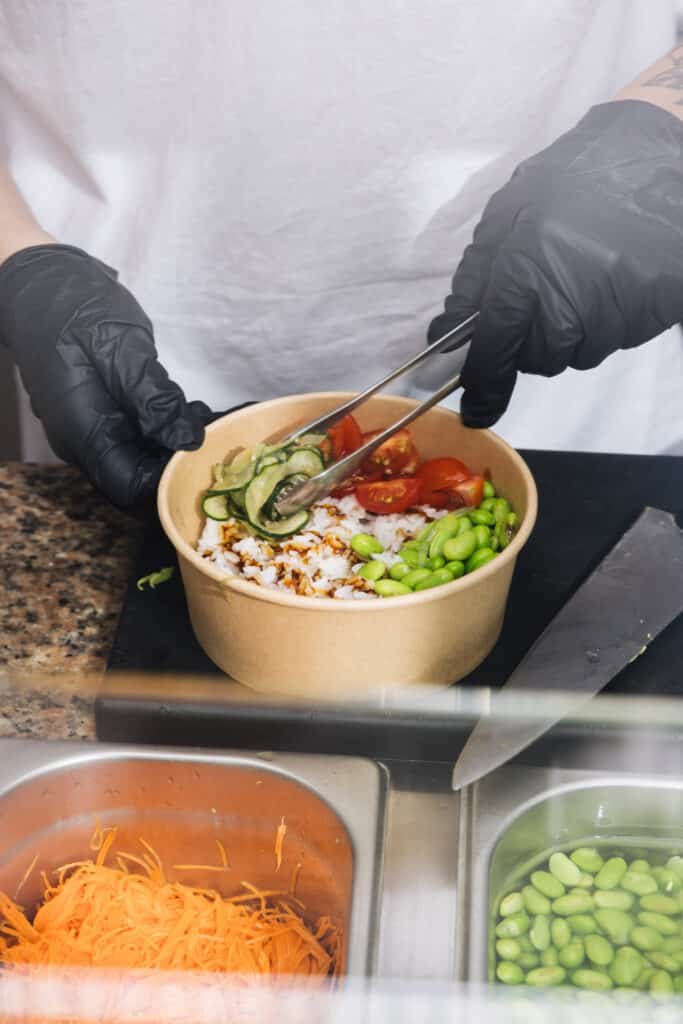 Una persona está preparando una comida en un tazón de papel marrón, utilizando unas pinzas para añadir rebanadas de aguacate a una variedad de ingredientes. En el tazón se puede ver una mezcla de arroz blanco y un tipo de grano tostado, tomates cherry cortados por la mitad, y edamames. La persona lleva guantes negros y una camiseta blanca, aunque no se muestra su rostro ni la parte superior de su cuerpo. Delante del tazón, hay un cuchillo de chef apoyado en la encimera, y a la izquierda, un recipiente de metal con zanahorias ralladas. El fondo es una cortina o pared de tela blanca, proporcionando un contraste simple que pone el énfasis en la acción de preparar la comida. En 4 pokes