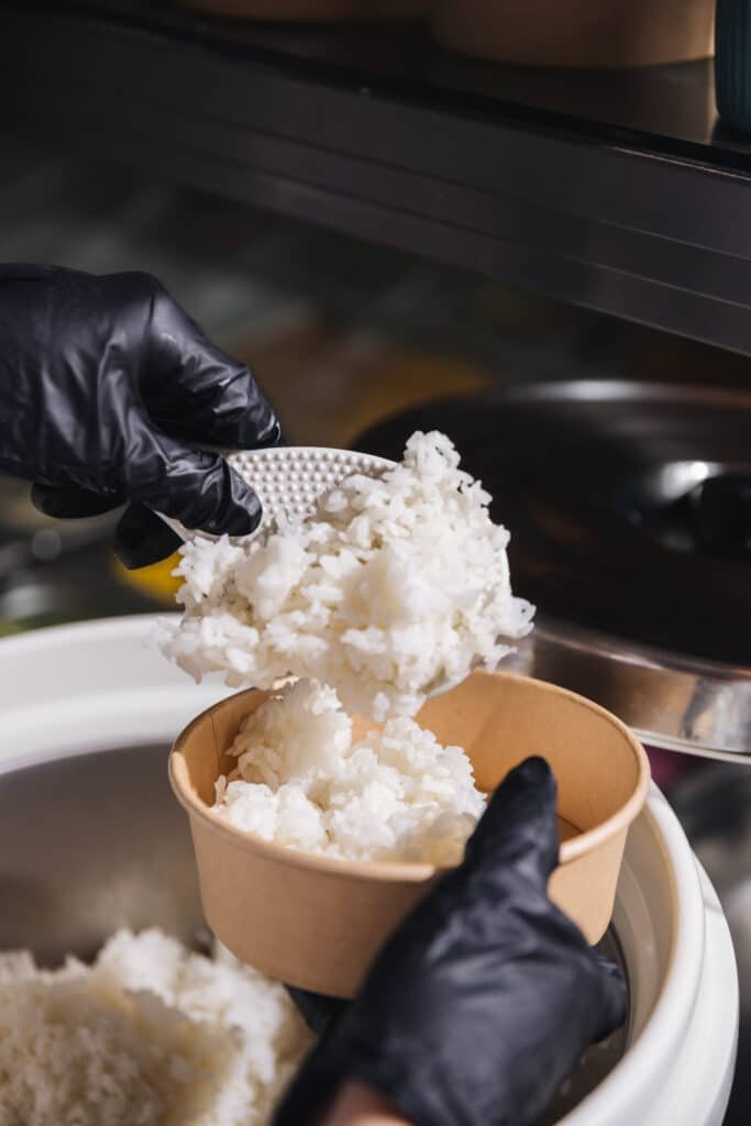 Una persona con guantes negros está sirviendo arroz blanco en un recipiente de papel marrón desde una olla de arroz. de 4 pokes